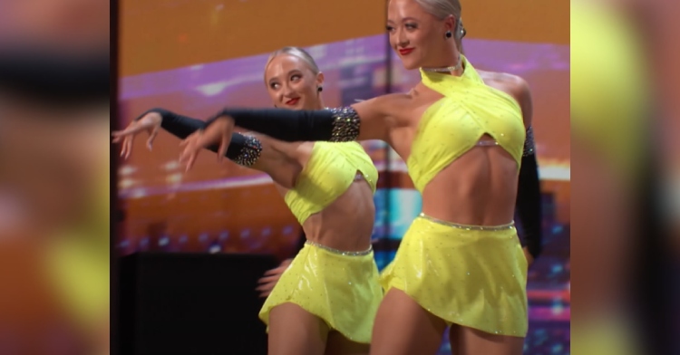 Twins smile as they dance in sync, each with an arm stretched out in front of them.