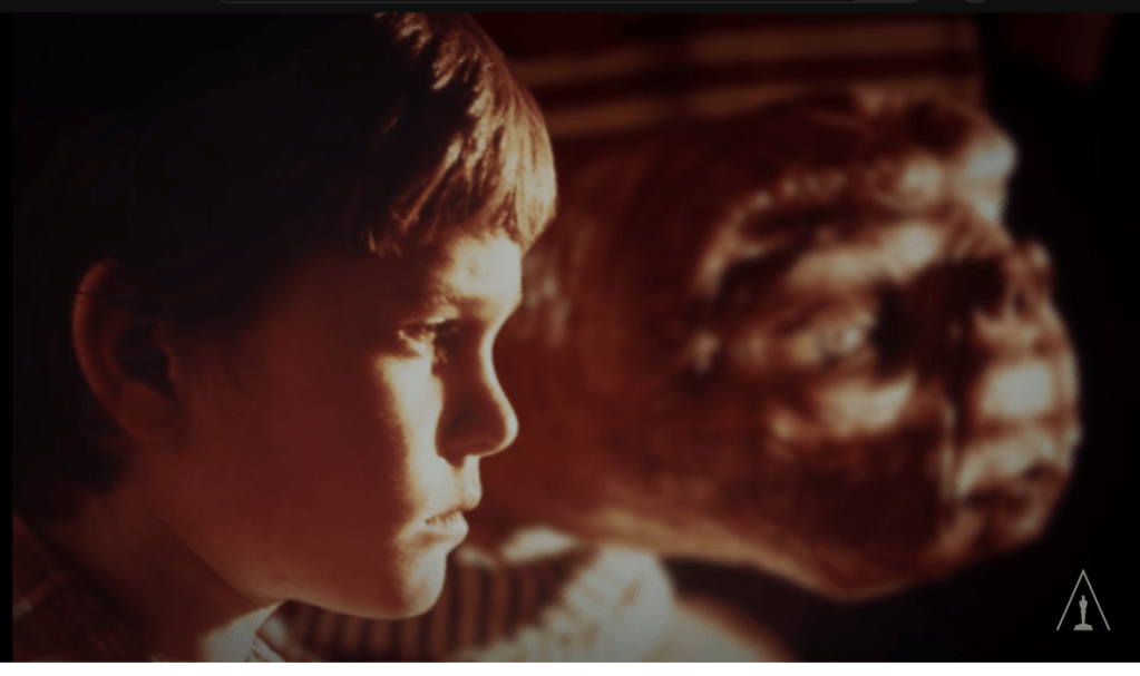 Henry Thomas as Elliott in "E.T." stares stoically out of a window in the distance with E.T. by his side doing the same. The shadows from the blinds can be seen on their faces with bits of light shining through.