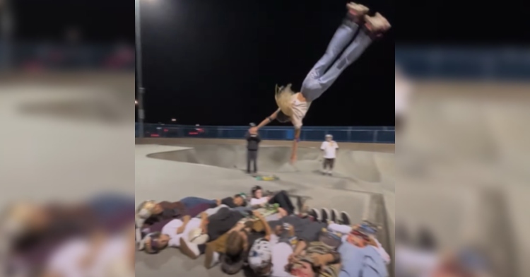 A teen, who is upside down, is midflight as she flips over 12 people laying on the ground while she's wearing roller skates.
