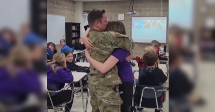 A man in a military uniform kisses the top of his daughter’s head as they hug in a classroom.