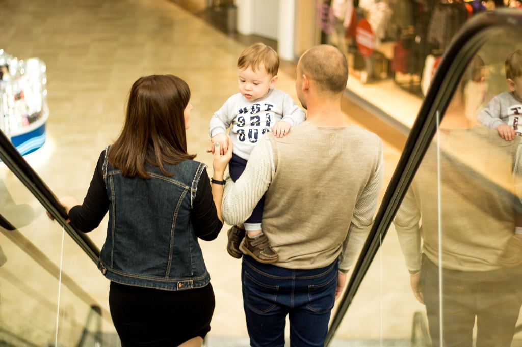 View from the back of a Mom, Dad, and toddler going down an escalator at a mall. The Dad is holding the kid in his arms while the Mom holds one of the kid's hands.