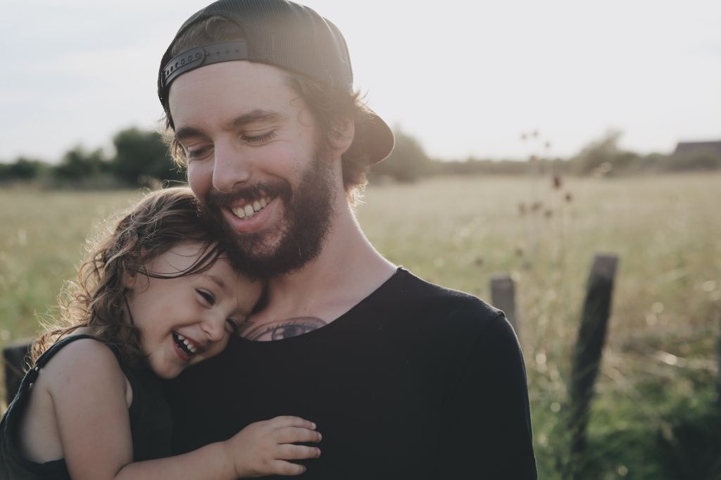 A man smiles and looks down as he holds his little girl in his arms. The little girl is smiling wide as she rests her head on his shoulder. They seem to be standing in a field but the background is out of focus.