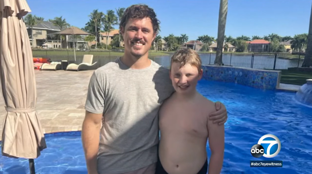 Jason Piquette smiles with one arm around Austen, who is also smiling and wearing his bathing suit. They're standing in front of a pool. 