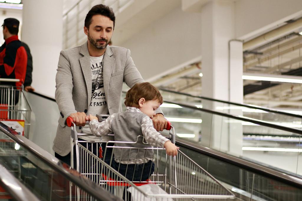 A dad looks down, seemingly deep in thought, as he pushes a shopping cart with his son in it. It seems they're on an escalator. 