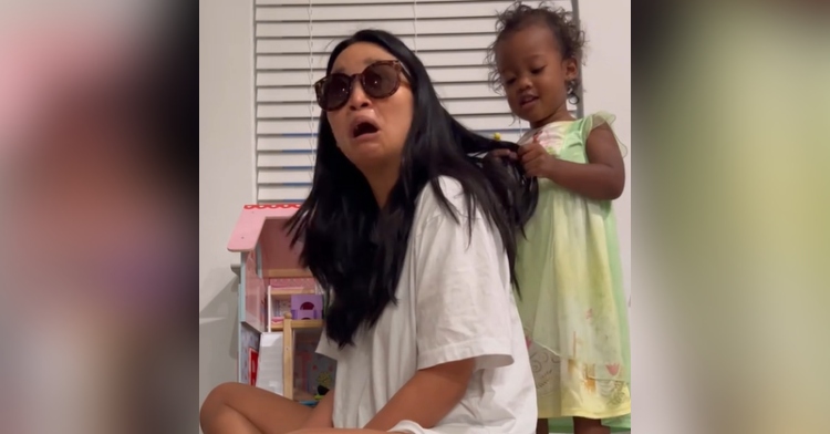 Mom sits on the floor while her toddler plays with her hair. Mom is wearing sunglasses and her mouth is agape from how shocked and moved she is by something her daughter said.