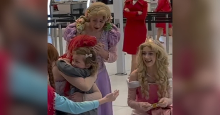 A woman dressed as Disney's Ariel hugs a little girl. Next to them are women dressed as Anna, Rapunzel, and Aurora. Rapunzel's mouth is agape, and she looks on the verge of tears.