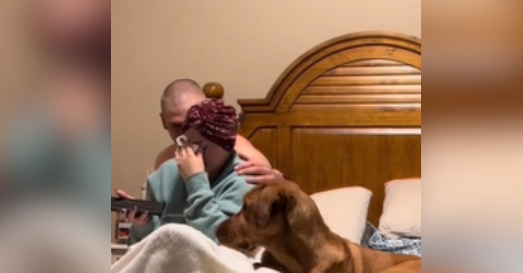 A woman sits in bed, face in her hands as she wipes her eyes with a tissue paper. With her other hand she holds a cell phone. A man sits behind her, hand on her shoulder for comfort. A large brown dog sits on the bed next to the woman.