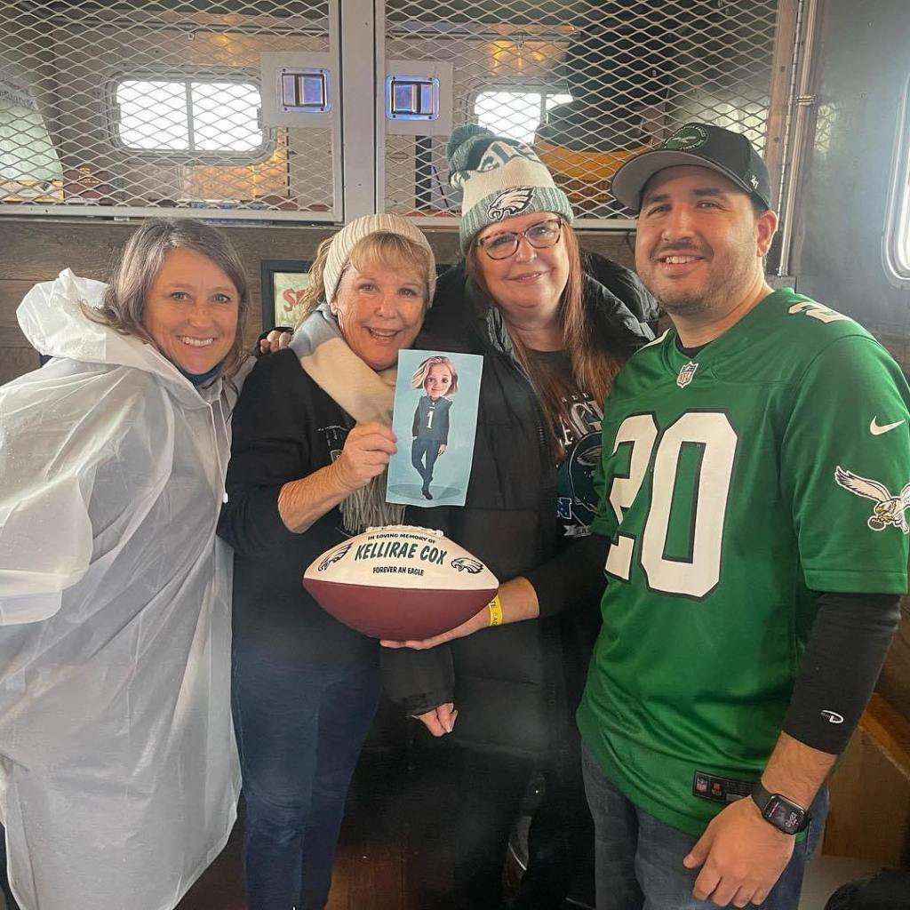 Rick smiles and poses for a photo with three of Kellirae's family members, including Suzanne McDonald. One of her family members holds up a cartoon photo of Kellirae and Rick holds the customized football with Kellirae's name.