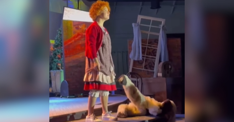 A little girl named Stella stands on a stage as she sings. She's playing the part of Little Annie. Next to her is a 6-year-old dressed as her dog, Sandy. The girl dressed as Sandy is falling off the stage, one foot straight in the air as she does.