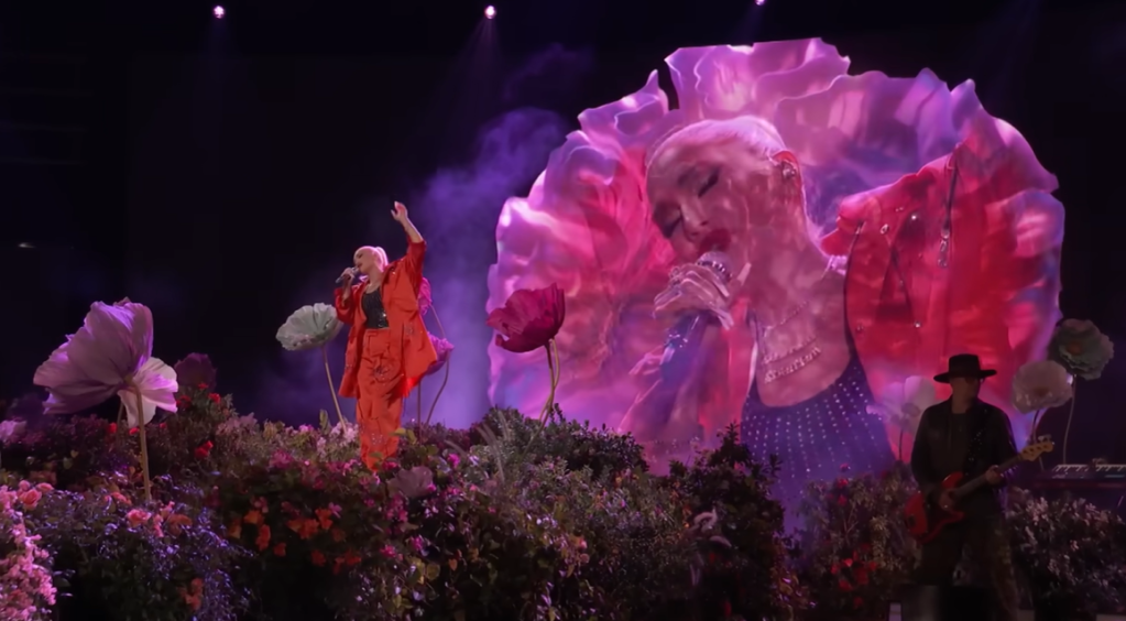 Gwen Stefani singing "True Babe" on "The Voice." As Stefani stands amidst the flora on stage, the screen behind her displays a close up of her face amidst wavy colors.