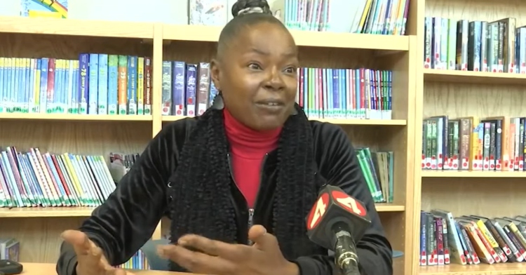 Teacher who saved a choking student, Tikiya Stewart, sits at a table in the school library and talks into a news mic.