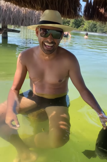 A man sits on a chair at a swim-up bar. He is wearing a swimsuit, hat and sunglasses. He is all smiles as he strokes the large, friendly fish.