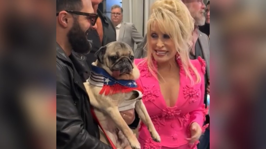 Dolly Parton smiles at the man holding Doug the Pug as the two of them meet.