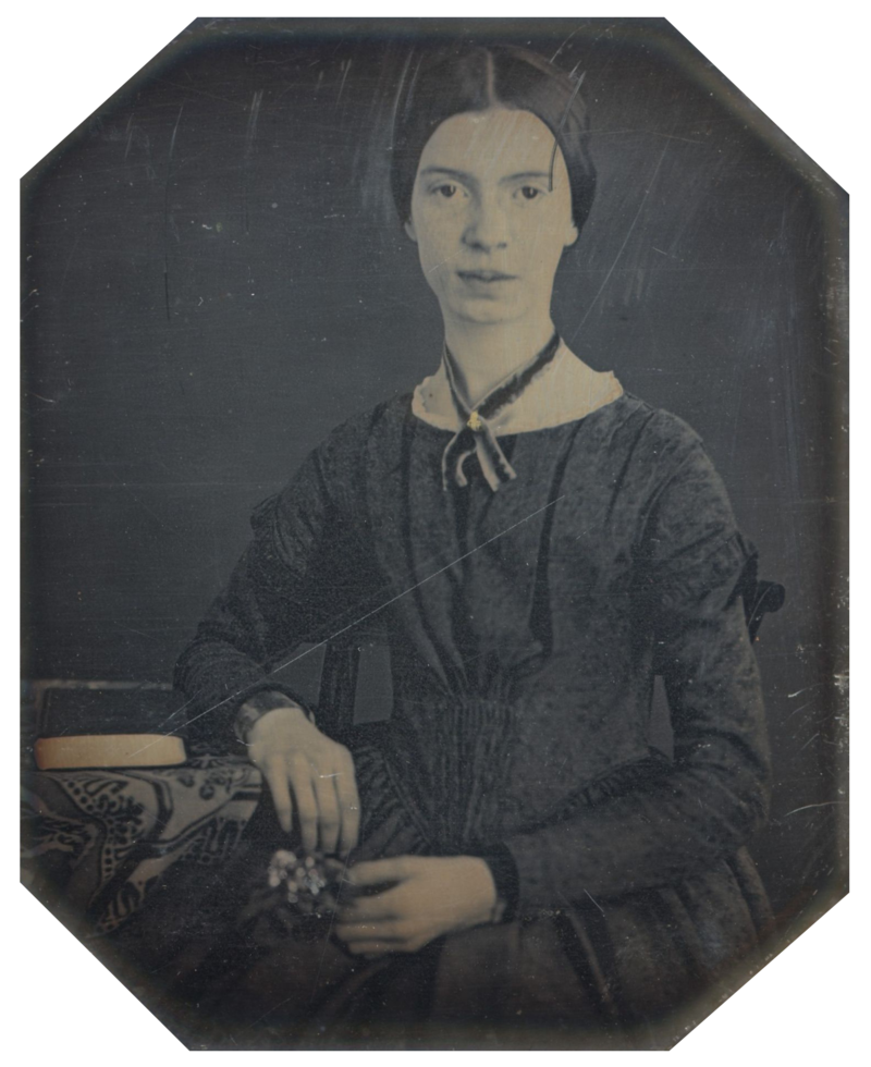 A black-and-white photo of Emily Dickinson. She has a slight smile on her face as she sits in a chair and rests her arm on a table that has a book.