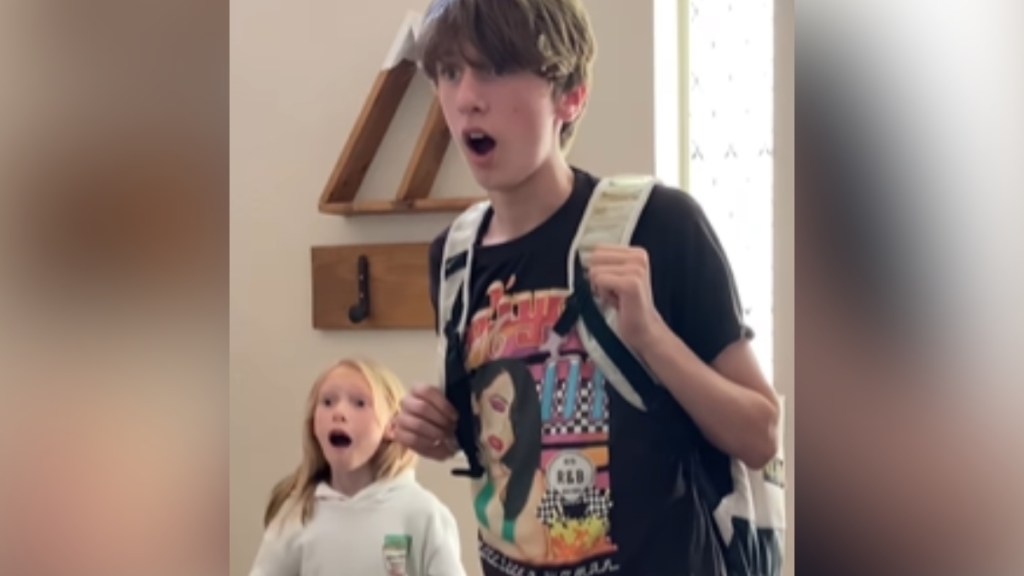 A teenage boy and a little girl looks shocked, mouths open, as they stand in the entrance of their home.