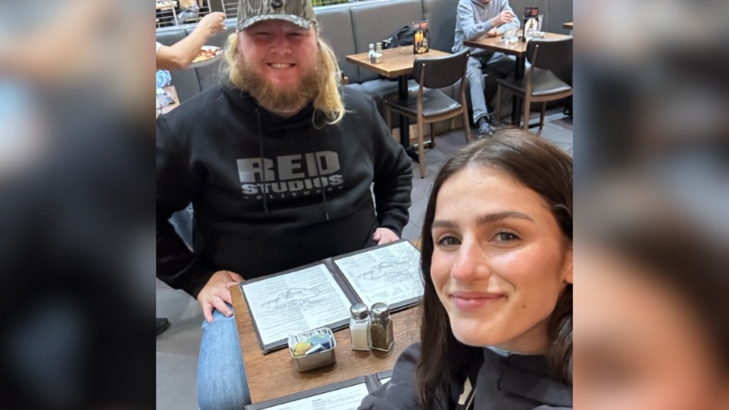 Abi Carter and Will Moseley smile as they sit at a restaurant table together. Abi is taking the photo.