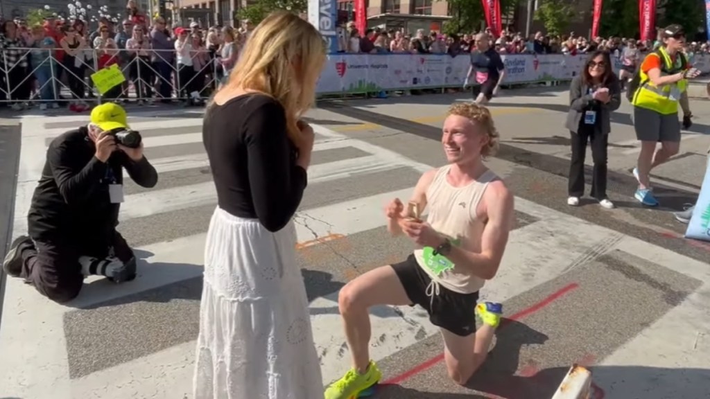 A marathon runner is bent on one knee, ring in hand to propose. A woman stands in front of him, back toward the camera. A photographer squats nearby, taking a photo of them
