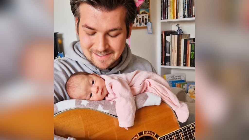 A man smiles as he sings and looks down. He's playing a guitar. Laying on top of his guitar is his newborn daughter.
