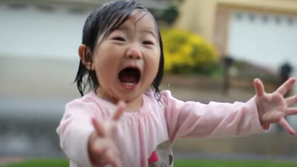 Close up of a little girl standing in the rain. Her arms are stretched out and her mouth is wide open from excitement.