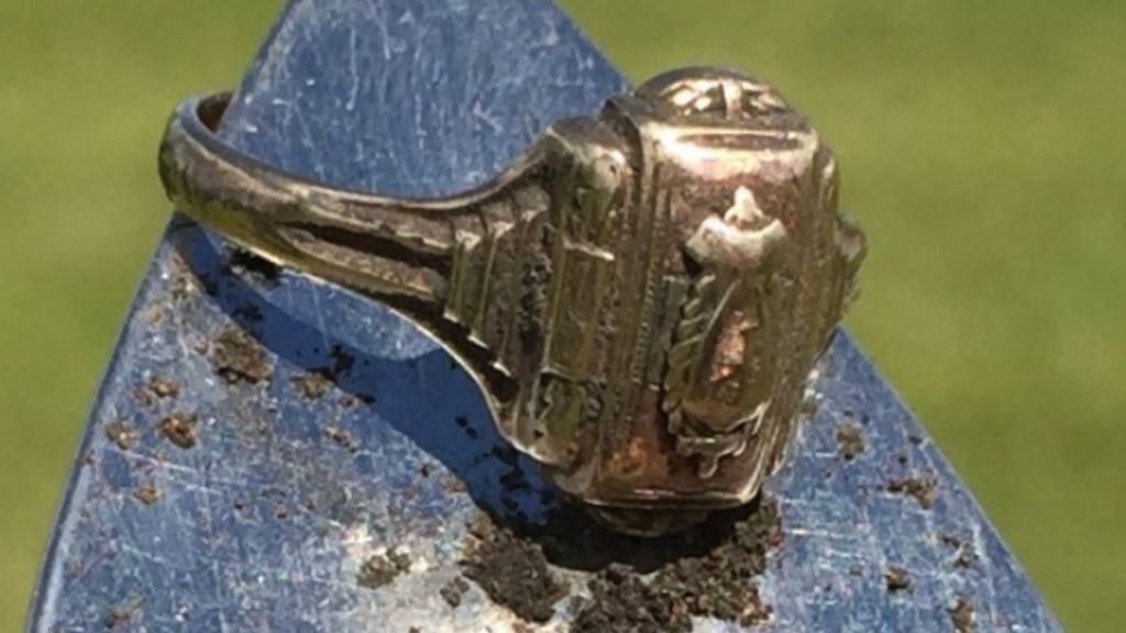 A gold class ring hanging from the tip of a dirt-covered shovel.