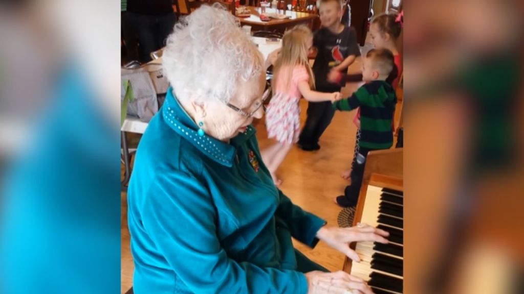 An elderly woman playing the piano while children dance in the background.