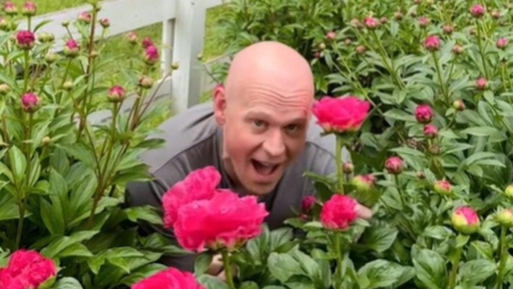 A smiling man crouches among the peonies in his garden.