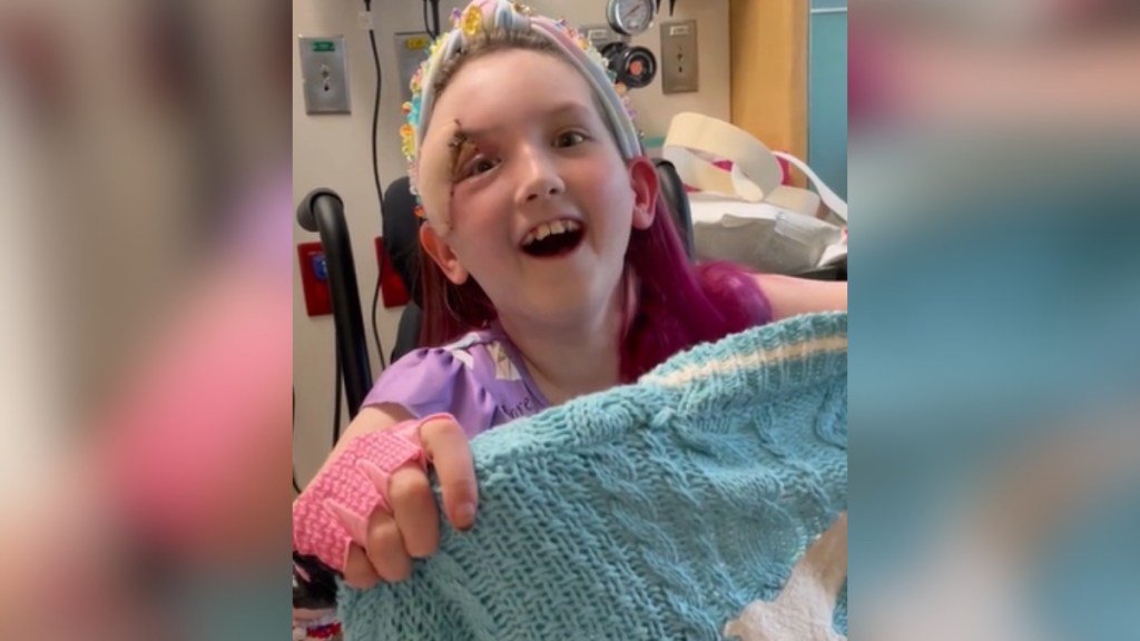 A little girl in a hospital holds up a "1989" cardigan from Taylor Swift. Her mouth is open from excitement.