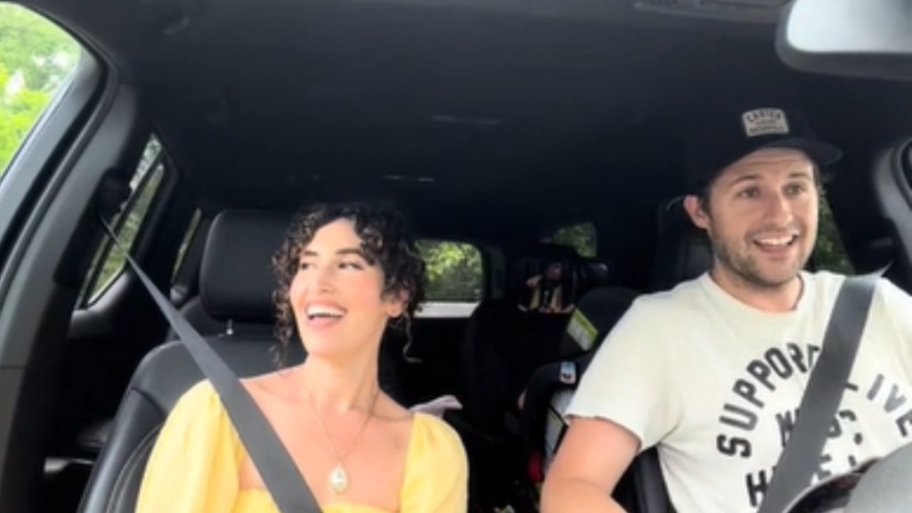 A woman and man smile as they sing during a car ride. He's driving and she's in the passenger seat.