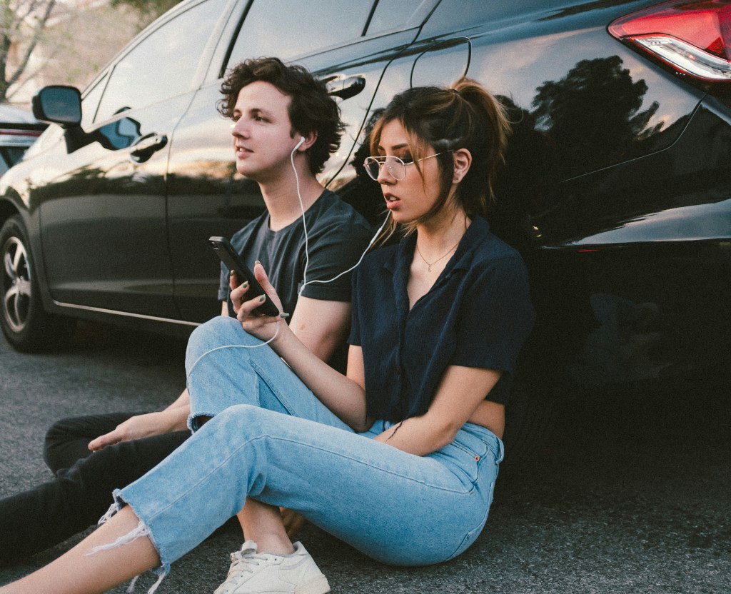 A man and woman share headphones connected to a phone the woman is holding. They're sitting on the ground and are leaning against a car. 