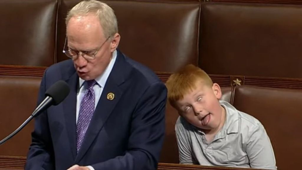 Image shows TN Rep. John Rose making a congressional speech while his son makes silly faces behind him.