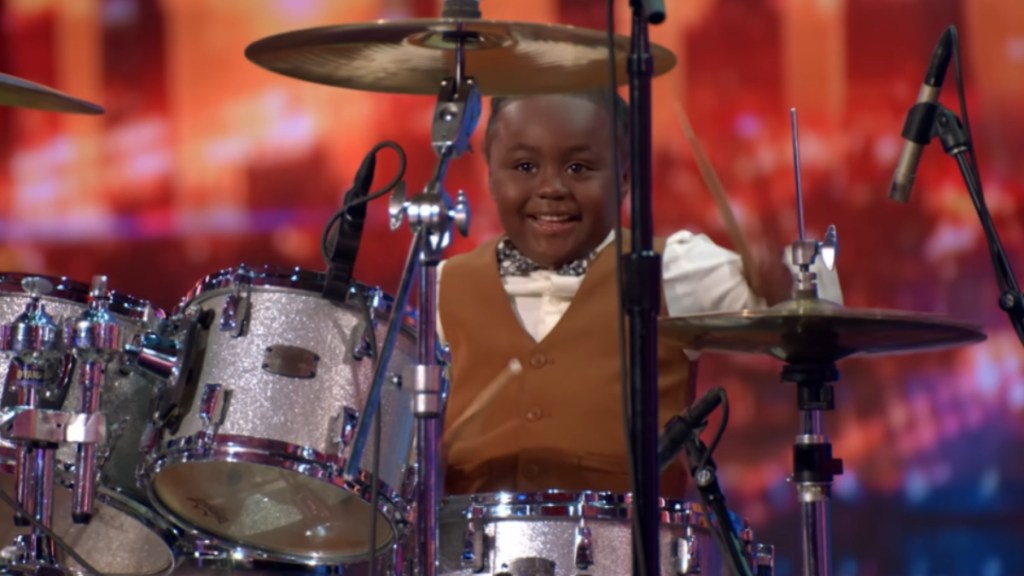 A little kid, Chrisyius Whitehead, smiles wide as he plays the drums.