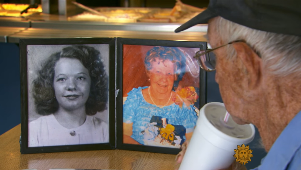 View from behind of a man sitting at a restaurant table, gazing at a two-photo frame with one containing a black-and-white photo of a young woman. The other shows the same woman, now elderly.