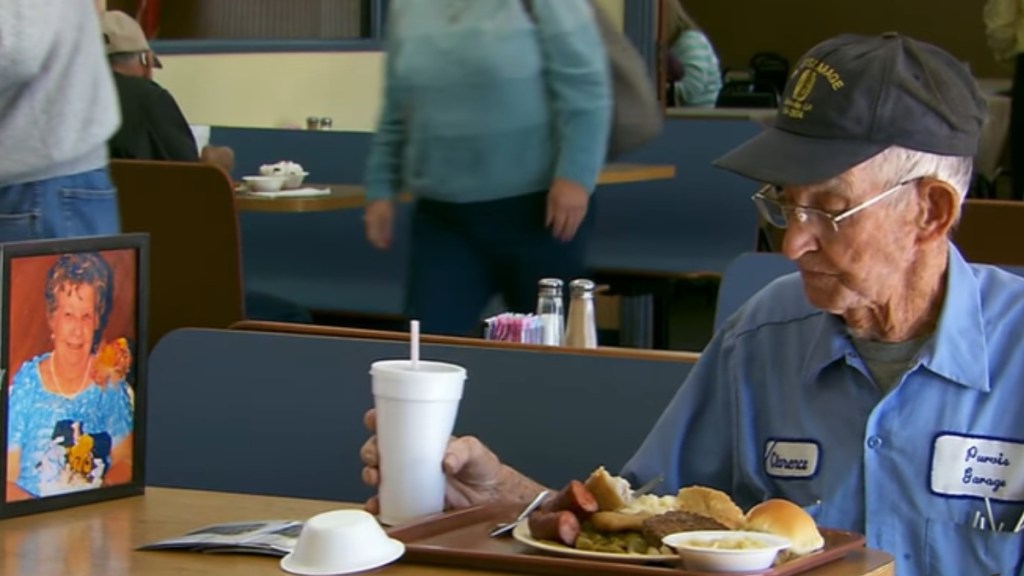 An elderly man sits at a restaurant booth, eating. With him at the table is a framed photo of an elderly woman