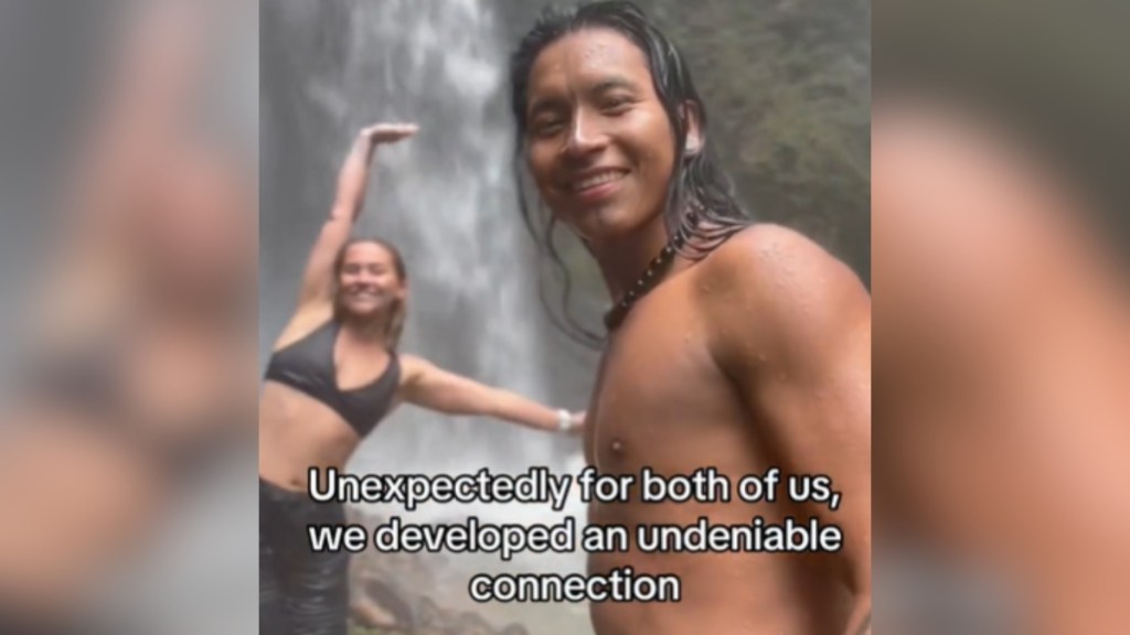 A man and woman pose for a photo. The man is closer to the camera and smiles. The woman is a bit farther away, arms in the air as she smiles. A waterfall is in the distance. Text on the image reads: Unexpectedly for both of us, we developed an undeniable connection