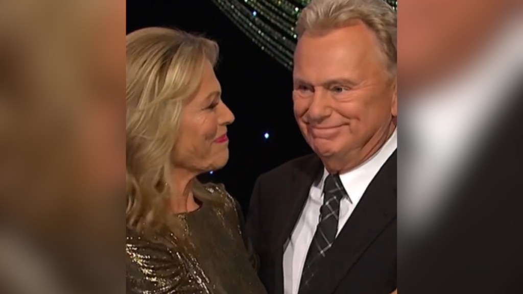 Pat Sajak smiles as Vanna White stands next to him, smiling at him.