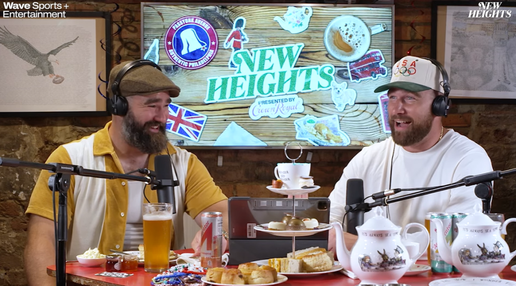 Jason and Travis Kelce sit at a table full of British foods and drinks for their podcast, New Heights. Jason smiles at his brother. Meanwhile, Travis has his mouth open wide, looking amazed as he talks.