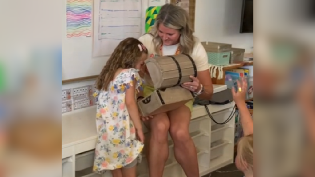 A teacher smiles as she holds open a treasure chest in her lap. A kindergartener peaks inside the treasure chest.