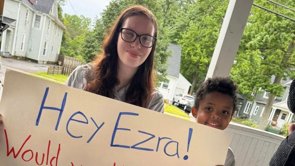 A young woman kneels next to a small child while holding a big sign.