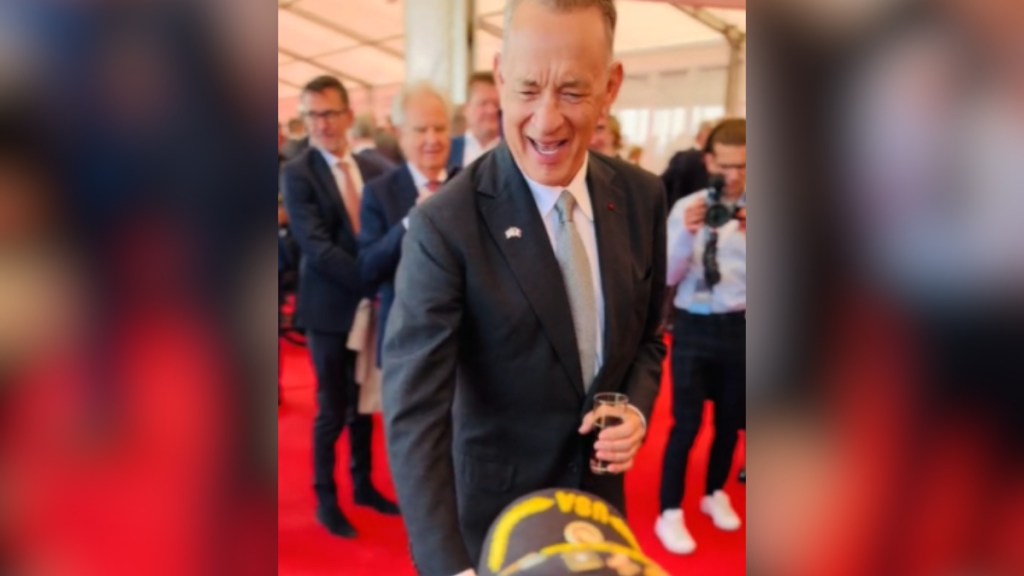 Tom Hanks smiles as he leans down to shake someone's hands - that person is sitting and we can only see the top of their hat.