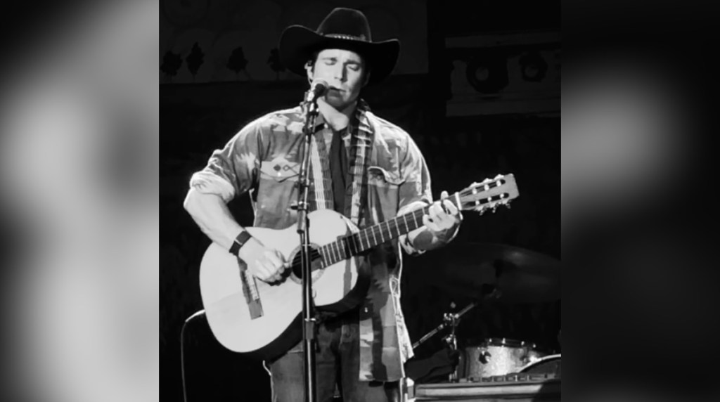 Black and white image of Lukas Nelson, Willie Nelson's son, singing on stage while playing a guitar. His eyes are closed as he sings and plays the guitar.