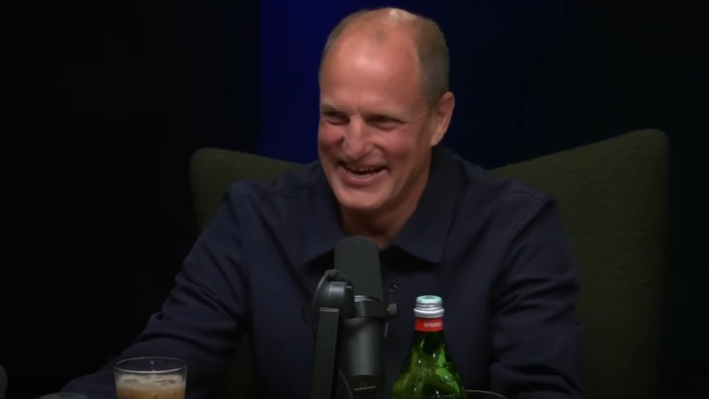 Woody Harrelson smiles as he sits behind a mic on a podcast