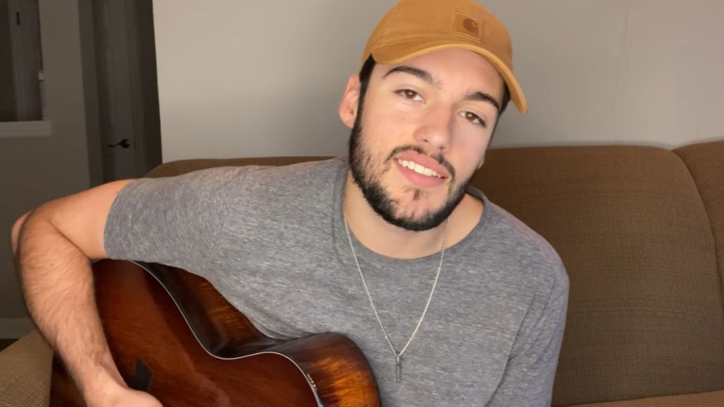 Garrett Jacobs smiles at the camera as he sits on a couch and sings while playing guitar