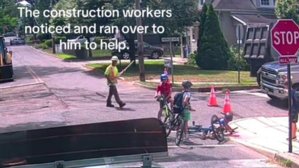 View from afar of kids gathered around a kid on the road who has fallen off his bike. Nearby, a construction worker is walking over to them. Text on the image reads: The construction workers noticed and ran over to him to help