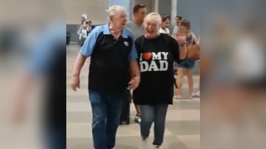 A dad and his adult daughter hold hands as they walk. She smiles wide, wearing a shirt that reads "I 'heart' my dad"