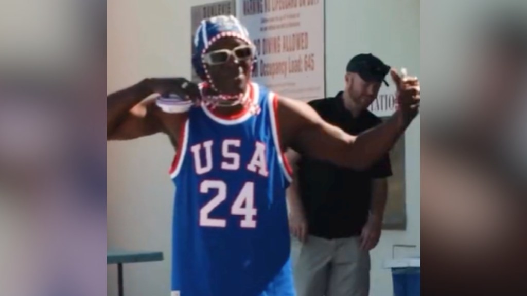 Flavor Flav holds up his clock necklace to show off his "USA 24" tank top. He's also wearing a water polo cap and glasses