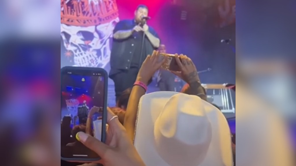 View from audience at a Jelly Roll concert. The singer stands on stage while one fan, who wears a large hat, holds up her phone with a message on it for him.