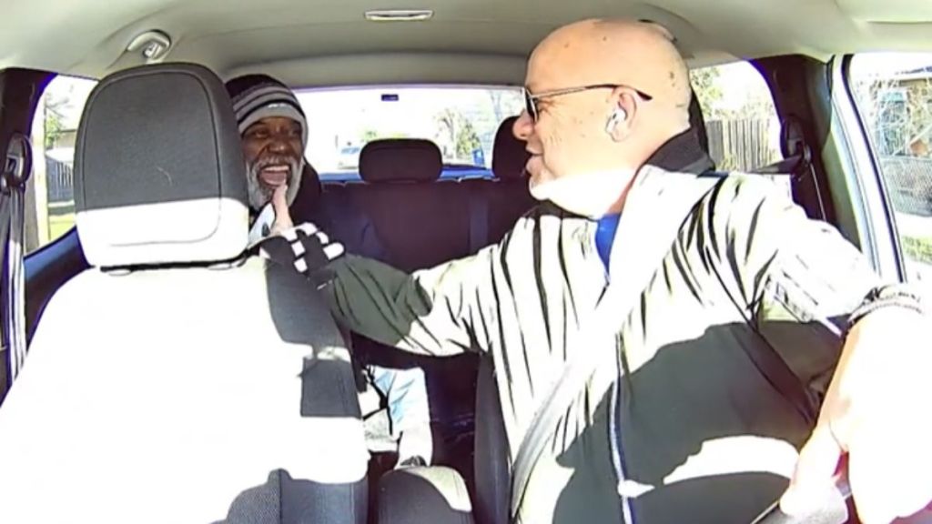 A driver turns around to shake hands with a smiling passenger.