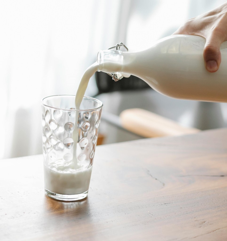 Close up of milk from a clear glass container being poured into a drinking glass sat on a table