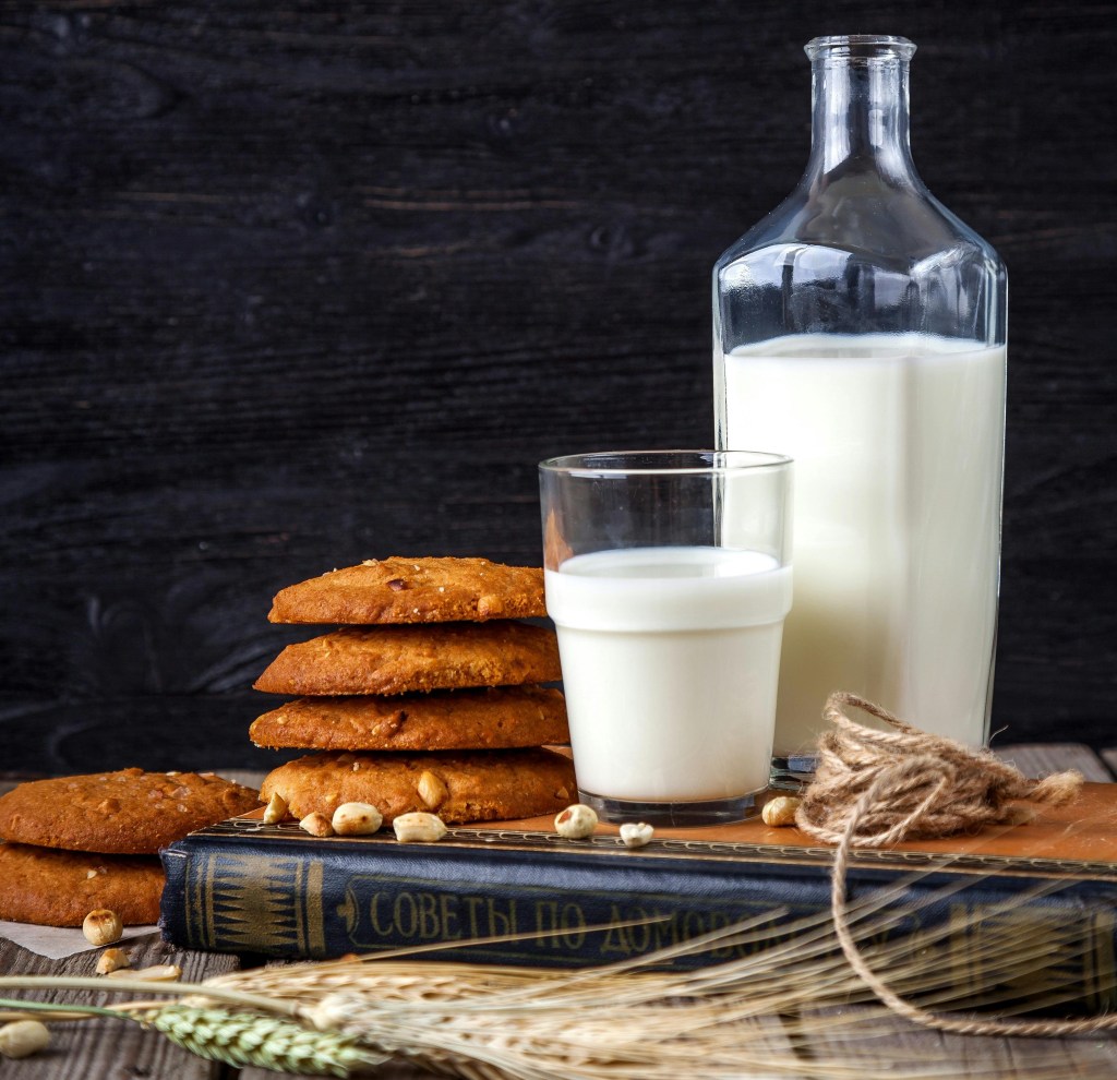 A glass container of milk sits next to a glass of milk and a stack of four cookies. All are placed on top of a book.