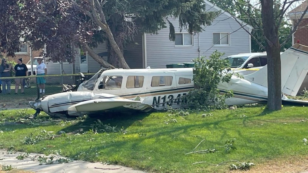 View of a small plane that has crashed in the front yard of a home. A few people stand at a distance being a fence nearby